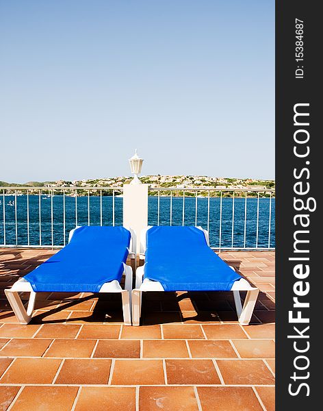 A horizontall image of two blue sun loungers on a terracota tiled patio in full sunshine with the sea in the background