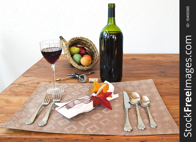 A bottle of wine, a glass of wine on a table with napkin, napkin ring with leaves, a cornucopia, silverware, placemat, and cork screw on a table. A bottle of wine, a glass of wine on a table with napkin, napkin ring with leaves, a cornucopia, silverware, placemat, and cork screw on a table.