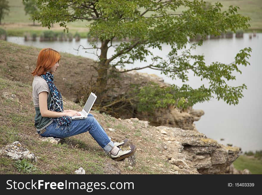 Young beautiful girl with laptop at rock near lake and tree.
