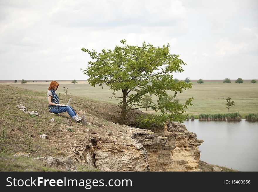 Girl with laptop at rock near lake and tree.