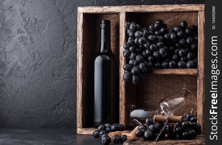 Luxury bottle of red wine and empty glasses with dark grapes inside vintage wooden box on black stone background. Corks and corkscrew on black board