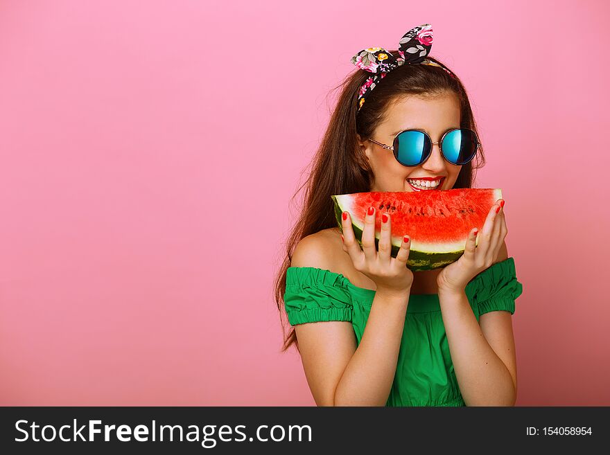 Portrait of a happy girl with long hair in green dress and sunglass, holding a slice of fresh watermelon in hand try to bite, over colorful pink background. Place for text. Portrait of a happy girl with long hair in green dress and sunglass, holding a slice of fresh watermelon in hand try to bite, over colorful pink background. Place for text.