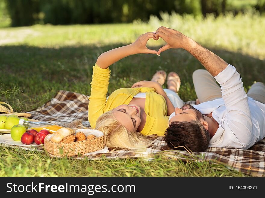 Happy couple in love enjoying day together, lying on plaid in park, making heart symbol