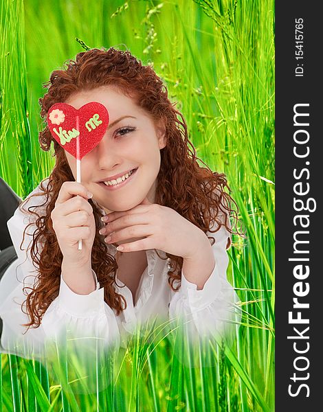 Good looking positive young caucasian girl with heartlike candy lying in green grass and smilng. Good looking positive young caucasian girl with heartlike candy lying in green grass and smilng