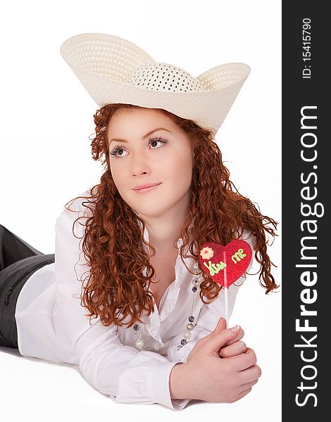 Young pretty caucasian girl wearing big hat and holding heartlike bon-bon candy dreaming about summer time and vacation isolated on white background. Young pretty caucasian girl wearing big hat and holding heartlike bon-bon candy dreaming about summer time and vacation isolated on white background