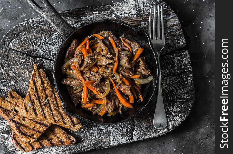 Slow cooker braised beef with onions and carrots in a pan on a dark background, top view.