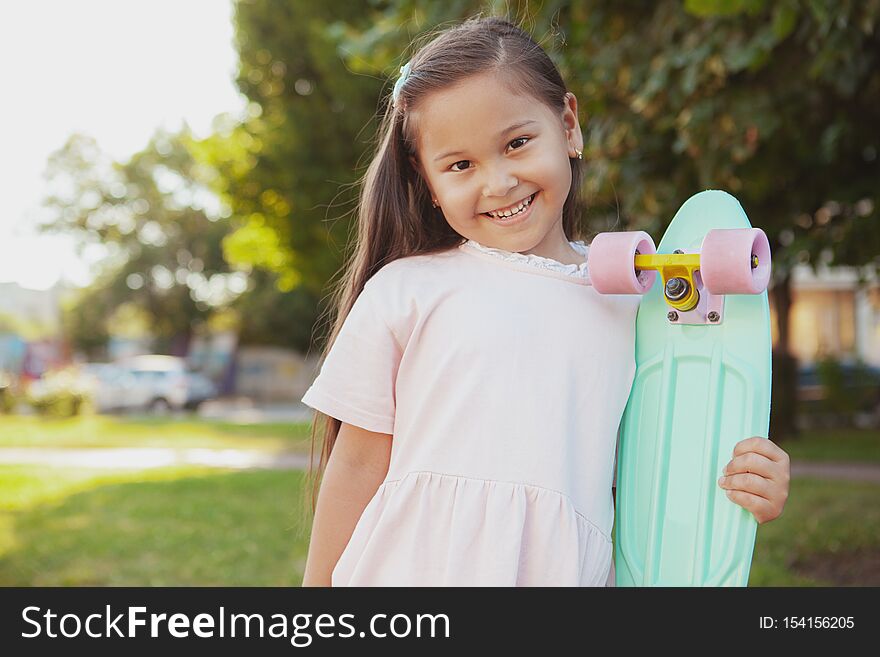 Charming little Asian girl smiling, holding her penny board at the park, copy space. Cheerful healthy little girl having fun outdoors, posing with her skateboard. Charming little Asian girl smiling, holding her penny board at the park, copy space. Cheerful healthy little girl having fun outdoors, posing with her skateboard