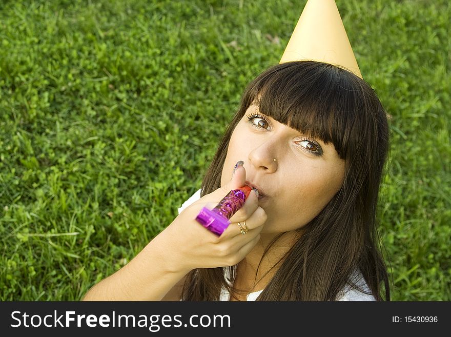 Beautiful girl sitting outdoors on grass, on his head festive hat in hand, whistle. Birthday party. Beautiful girl sitting outdoors on grass, on his head festive hat in hand, whistle. Birthday party