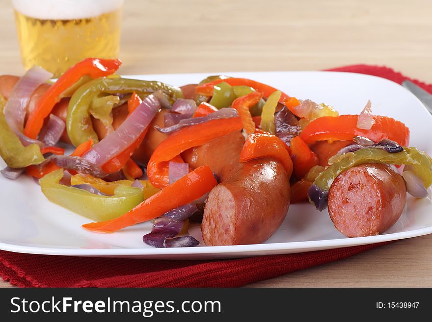 Two smoked sausages on a plate covered with red and green bell peppers and onions with a beer in the background. Two smoked sausages on a plate covered with red and green bell peppers and onions with a beer in the background