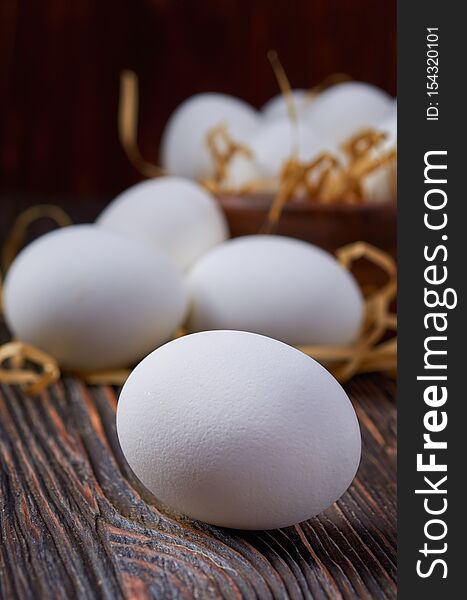White eggs on a wooden table. In the background are eggs in a wooden bowl and on paper straw. Close up. Low key. White eggs on a wooden table. In the background are eggs in a wooden bowl and on paper straw. Close up. Low key