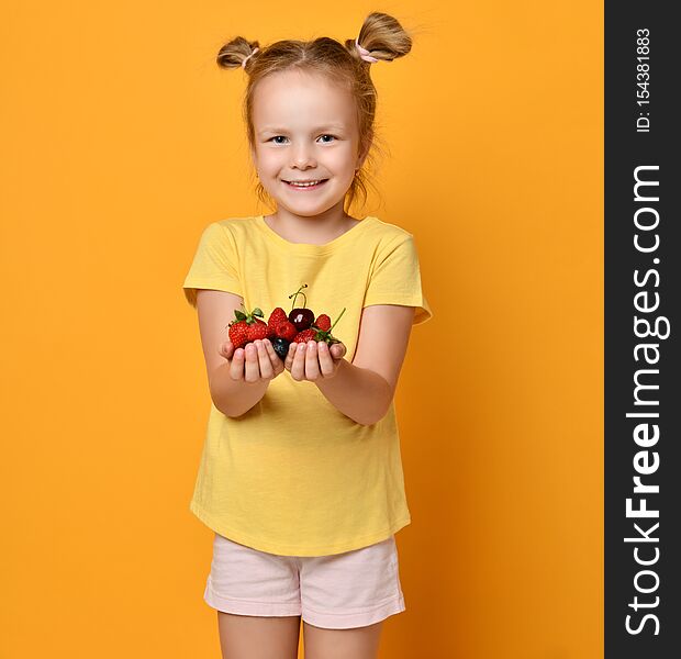 Baby girl kid blonde with funny buns demonstrates a handful of berries fruits in her palms hands and smiles satisfied with her harvest on yellow  background. Baby girl kid blonde with funny buns demonstrates a handful of berries fruits in her palms hands and smiles satisfied with her harvest on yellow  background