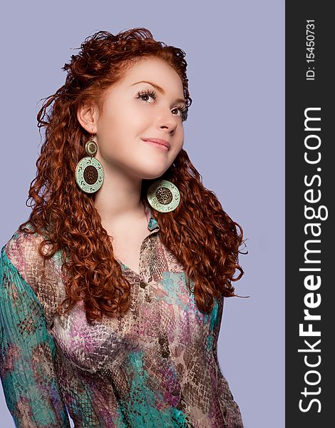 Relaxing and dreaming sensual red haired caucasian girl with positive and calm facial expression wearing extravagant round earrings and bright multicolored shirt standing isolated over blue background. Relaxing and dreaming sensual red haired caucasian girl with positive and calm facial expression wearing extravagant round earrings and bright multicolored shirt standing isolated over blue background
