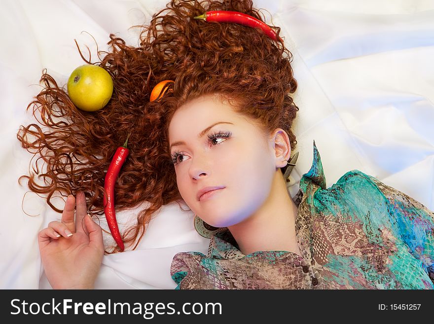 Multicolored fruits and chili pepper laying in hair of young caucasian girl lying on white linen with calm facial expression. Multicolored fruits and chili pepper laying in hair of young caucasian girl lying on white linen with calm facial expression