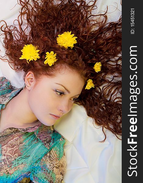 Extravagant and bright looking caucasian red haired beautiful girl having yellow flowers interlaced in her hair lying over white riffled linen with calm and dreaming facial expression. Extravagant and bright looking caucasian red haired beautiful girl having yellow flowers interlaced in her hair lying over white riffled linen with calm and dreaming facial expression