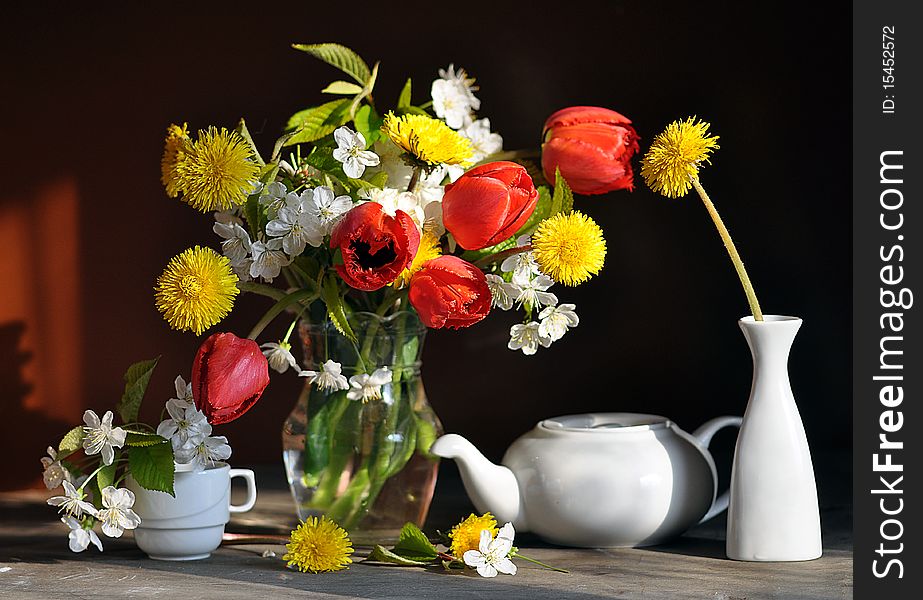 tulips, dandelions and branch of flowering cherry, stand in a glass vase, alongside tea-pot and cup. tulips, dandelions and branch of flowering cherry, stand in a glass vase, alongside tea-pot and cup