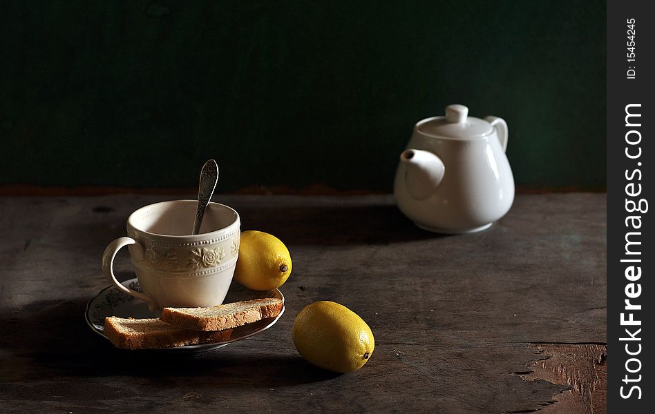 A cup stands on an old table, two lemons, bread, lie, a bit farther a tea-pot stands from a cup. A cup stands on an old table, two lemons, bread, lie, a bit farther a tea-pot stands from a cup