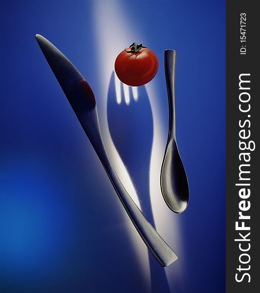 Tomato with knife and spoon on blue background with silhouetted fork underneath. Tomato with knife and spoon on blue background with silhouetted fork underneath