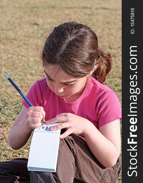 Cute little girl with pigtails writes in the notepad sitting on the grass outdoor. Cute little girl with pigtails writes in the notepad sitting on the grass outdoor