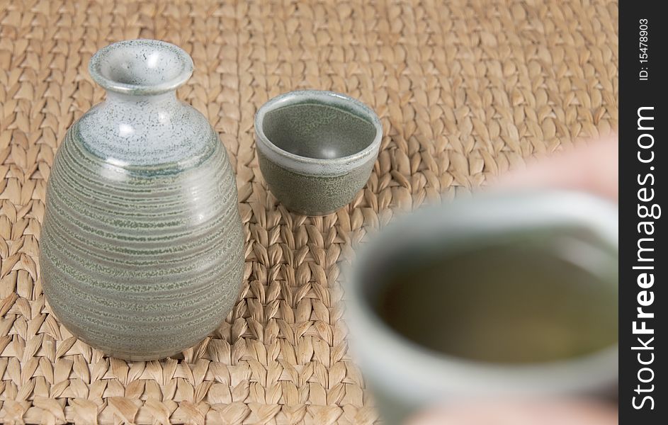 A green pottery tea set on woven bamboo mat with one cup raised to drink. A green pottery tea set on woven bamboo mat with one cup raised to drink