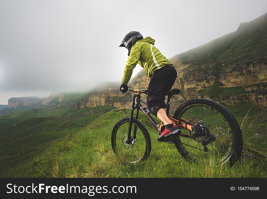 Aged male athlete in helmet and mask rides a mountain bike on a grassy slope against the background of plateau rocks and