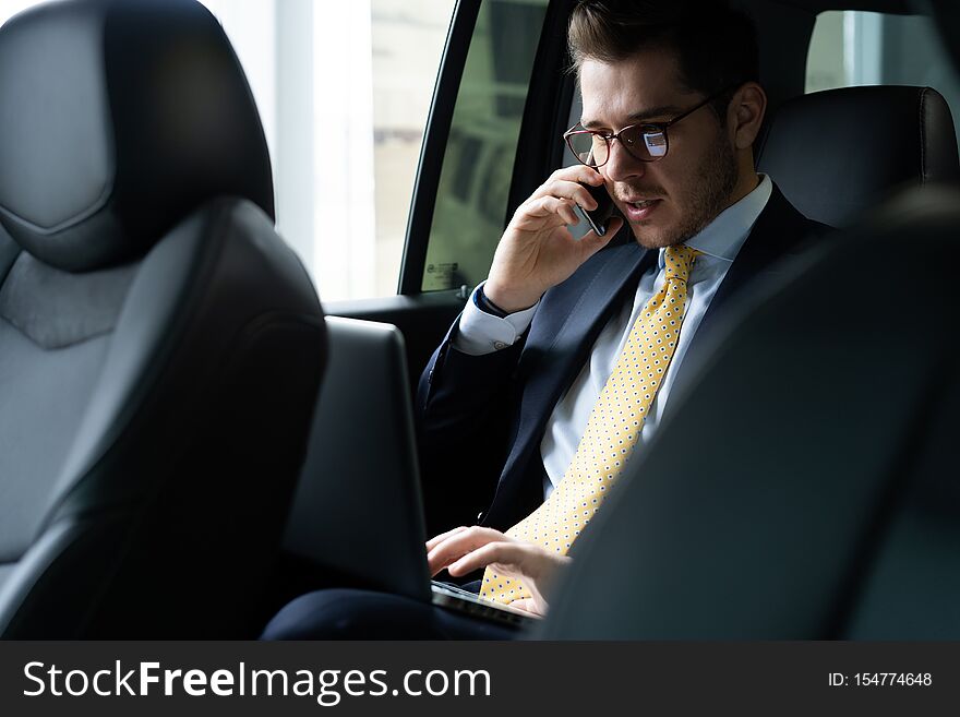Young businessman sitting on back seat of the car, while his chauffeur is driving automobile.