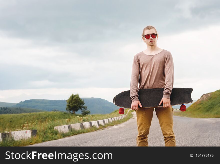 A stylish young man standing along a winding mountain road with a skate or longboard in his hands the evening after