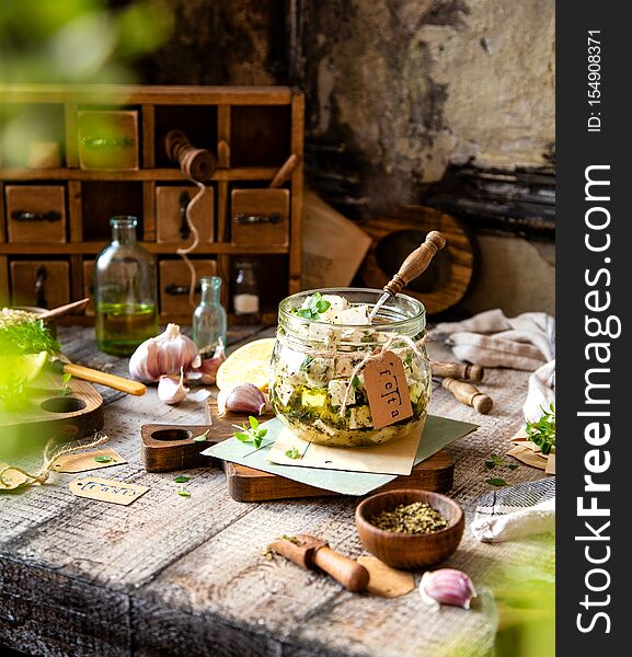 Homemade marinated diced cheese feta in glass jar with herbs, garlic, basil on wooden board with old paper on grey wood table with bottle of oil, lemon, leaves, napkin opposite brown chest of drawers