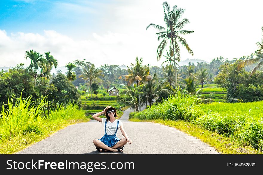 Tropical portrait of young happy woman with straw hat on a road with coconut palms and tropical trees. Bali island. Indonesia.