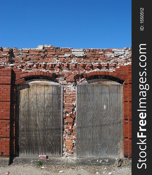 Description: Industrial ruins of two boarded-up doors framed by bricks.  There is a blue sky in the top third of the photo.  The top portion of the bricks are crumbling. Description: Industrial ruins of two boarded-up doors framed by bricks.  There is a blue sky in the top third of the photo.  The top portion of the bricks are crumbling.