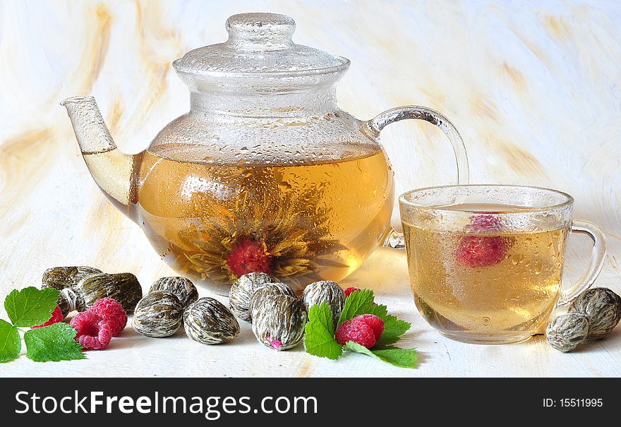 Tea is brewed in a glass tea-pot, alongside cup, knitted tea and raspberry. Tea is brewed in a glass tea-pot, alongside cup, knitted tea and raspberry