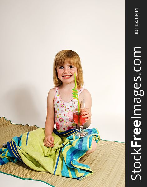 A vertical image of a lovely young girl in bathing costume holding a drink and sitting on a beach mat as she plays at being at the seaside,isolated against a plain background. A vertical image of a lovely young girl in bathing costume holding a drink and sitting on a beach mat as she plays at being at the seaside,isolated against a plain background