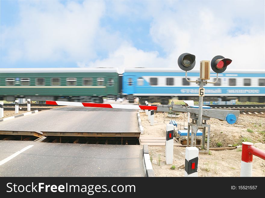 The image of railway crossing and the train. Focus is under the traffic light. Train us blurred.