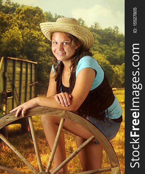 A happy young teen wearing cowgirl gear while in a field leaning on a wooden wagon wheel. A happy young teen wearing cowgirl gear while in a field leaning on a wooden wagon wheel.