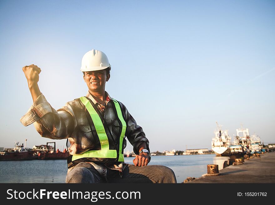 Male engineer working at the harbor wearing a white hat, raising both hands