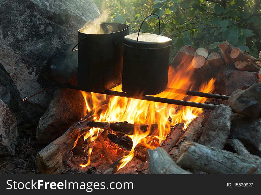 Two tourist kettles over campfire around rock with firewood