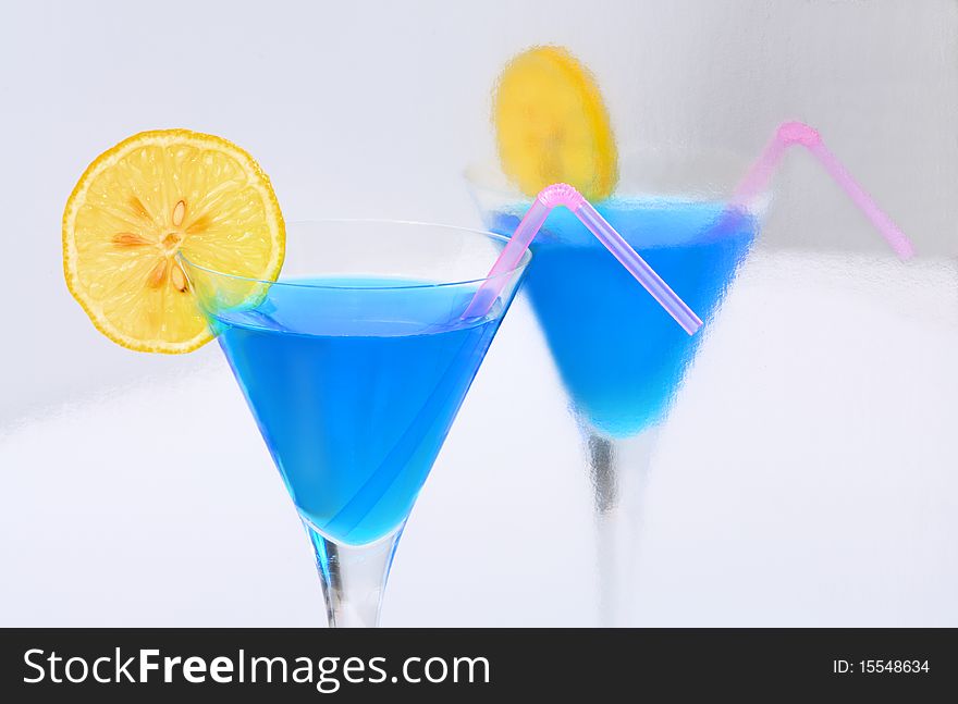 A blue cocktail with a lemon & a straw reflected in a silvery surface. A blue cocktail with a lemon & a straw reflected in a silvery surface.