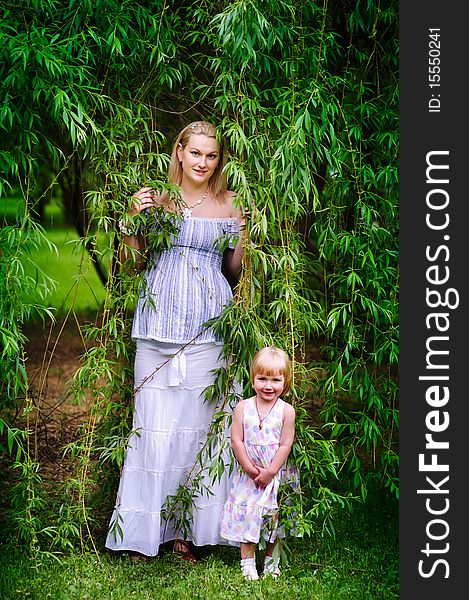 Beautiful pregnant woman and her adorable baby daughter stand in the willow garden. Beautiful pregnant woman and her adorable baby daughter stand in the willow garden