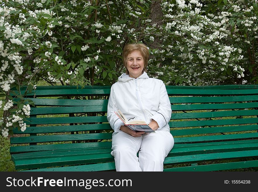 Elderly woman with a book in hands. Sits on a bench in a garden
Smiles and looks in a photographic camera