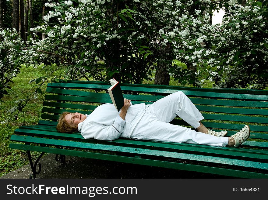 Ŭderly woman reads a book lying on a bench in a garden near a flowering bush