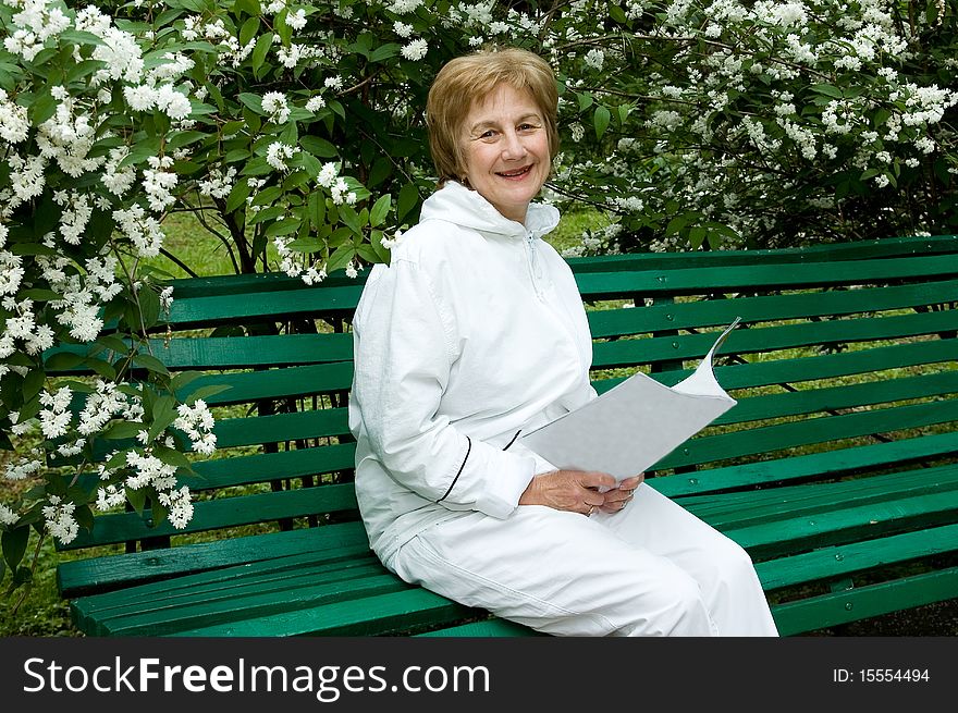 Elderly woman with a illustrated magazine in hands. Sits on a bench in a garden
Smiles and looks in a photographic camera