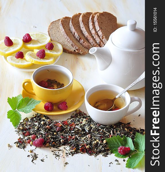 Two cups herbal tea is brewed in which, alongside tea-pot, lemon, bread and raspberry