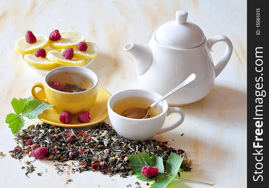 Two cups herbal tea is brewed in which, alongside tea-pot, lemon, and raspberry