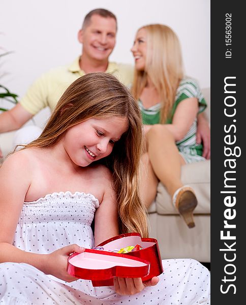 Surprised young cute girl holding her gift with parents in the background. Surprised young cute girl holding her gift with parents in the background