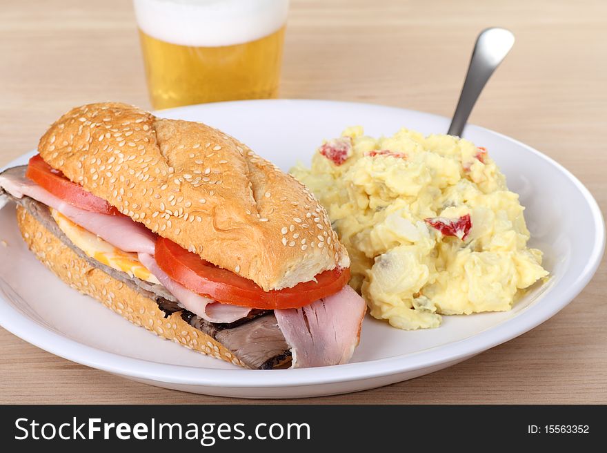 Ham and roast beef sub with tomatoes and cheese on a sesame seed bun with potato salad and a beer. Ham and roast beef sub with tomatoes and cheese on a sesame seed bun with potato salad and a beer