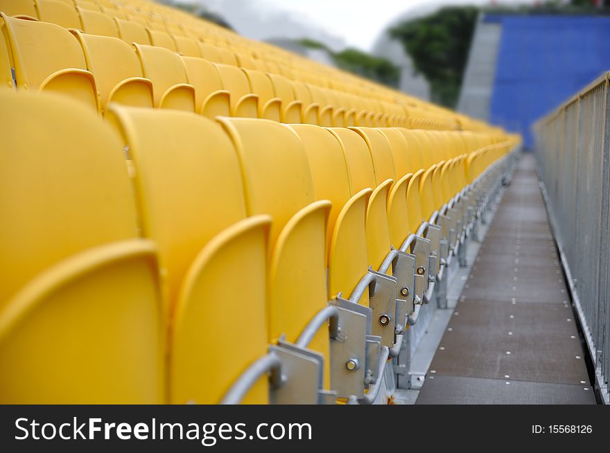 Front row of a section of folded yellow spectator seats with fence and a walkway leading to a section of blue seats. Front row of a section of folded yellow spectator seats with fence and a walkway leading to a section of blue seats.