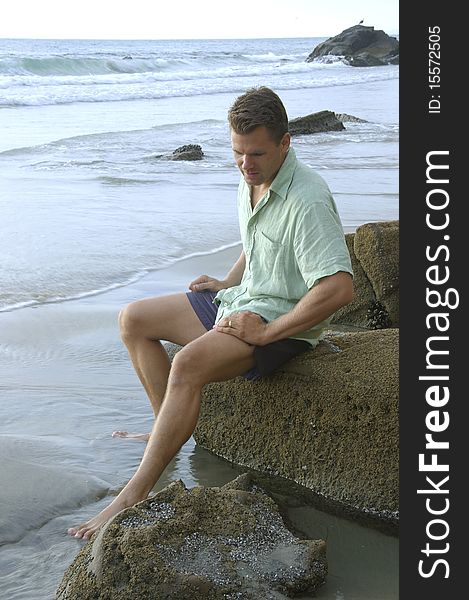 Man sits on rock while touching beach water with feet. Man sits on rock while touching beach water with feet