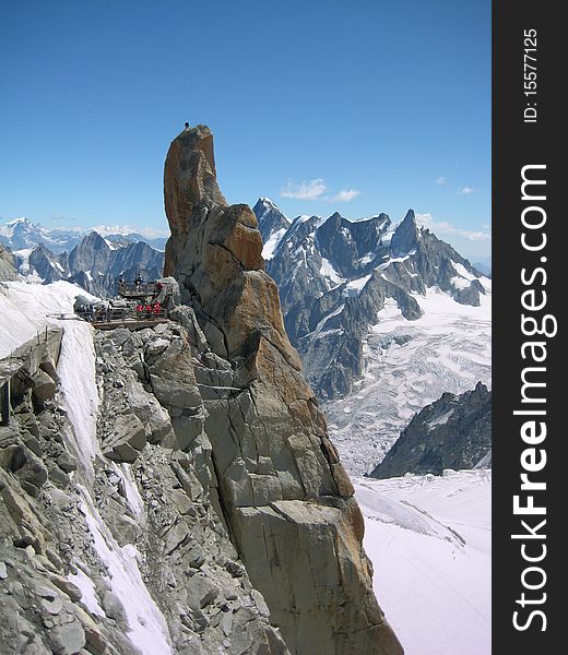 Climber standing on the exposed summit pinnacle of the Aiguille du Midi Face Sud multipitch climb, watched by tourists at the cablecar station. Climber standing on the exposed summit pinnacle of the Aiguille du Midi Face Sud multipitch climb, watched by tourists at the cablecar station.