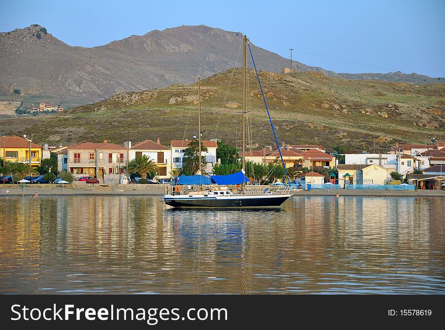 Dark blue sailboat reflected in calm sea water at sunset, in front of lovely small fishing village