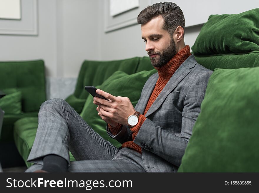 Focused handsome man in formal wear sitting on green sofa and using smartphone