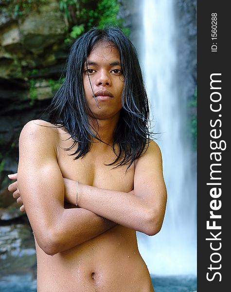 Asian exotic long-haired boy in front of a tropical pristine waterfall, after taking a dip in the whirlpool, shivering and covered with droplets. Asian exotic long-haired boy in front of a tropical pristine waterfall, after taking a dip in the whirlpool, shivering and covered with droplets.